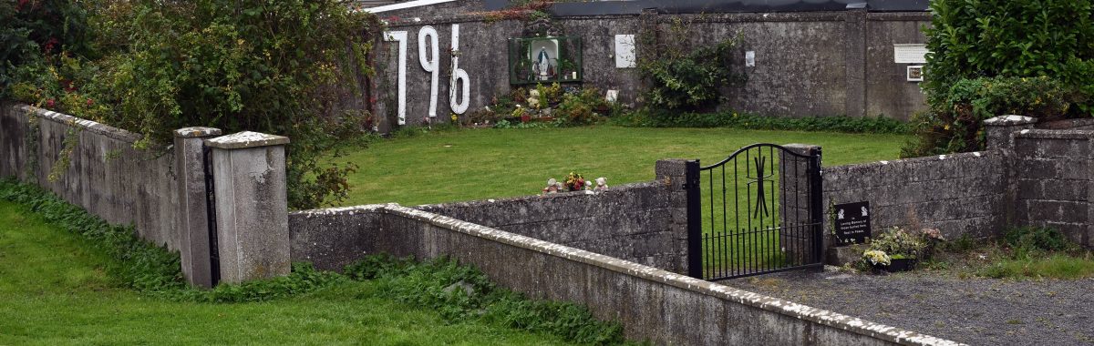 Image of grey walled Memorial Garden at site of the former Mother and Baby Home in Tuam, large white letters 796 on the back wall, black iron gate and memorial plaque at the entrance, flowers and statue